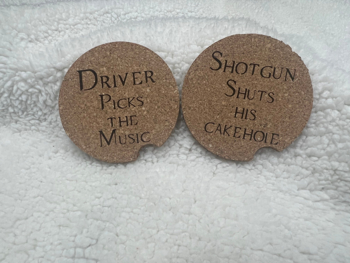The Set of 2 cork car coasters by MOON AND SKYE CREATIONS LLC adds a personalized touch to any drive with the phrases Driver picks the music and Shotgun shuts his cakehole on a textured white surface.