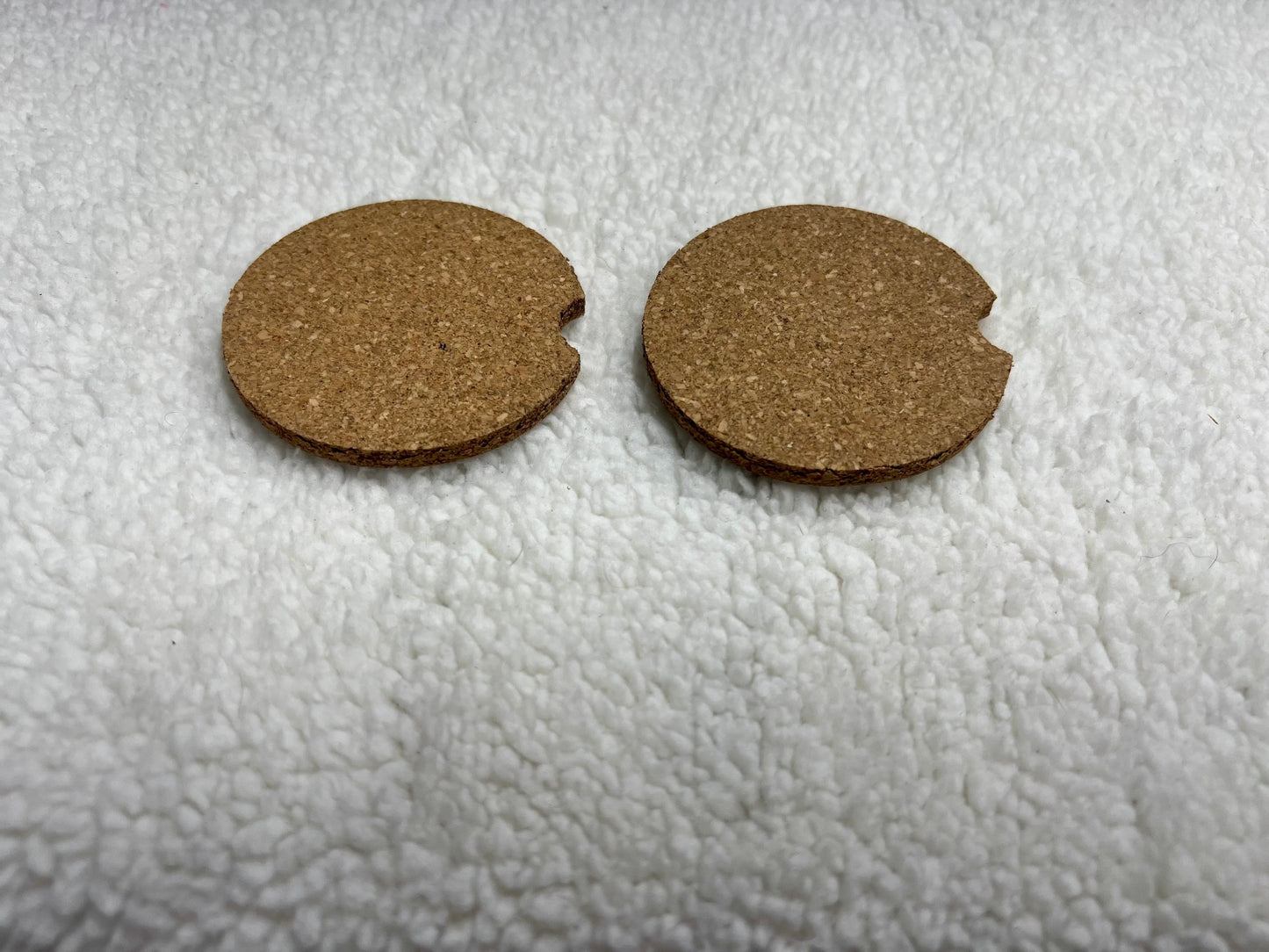 The Set of 2 cork car coasters by MOON AND SKYE CREATIONS LLC, in natural brown, features identical circular designs with notches, resting stylishly on a soft white textured surface for a personalized touch.