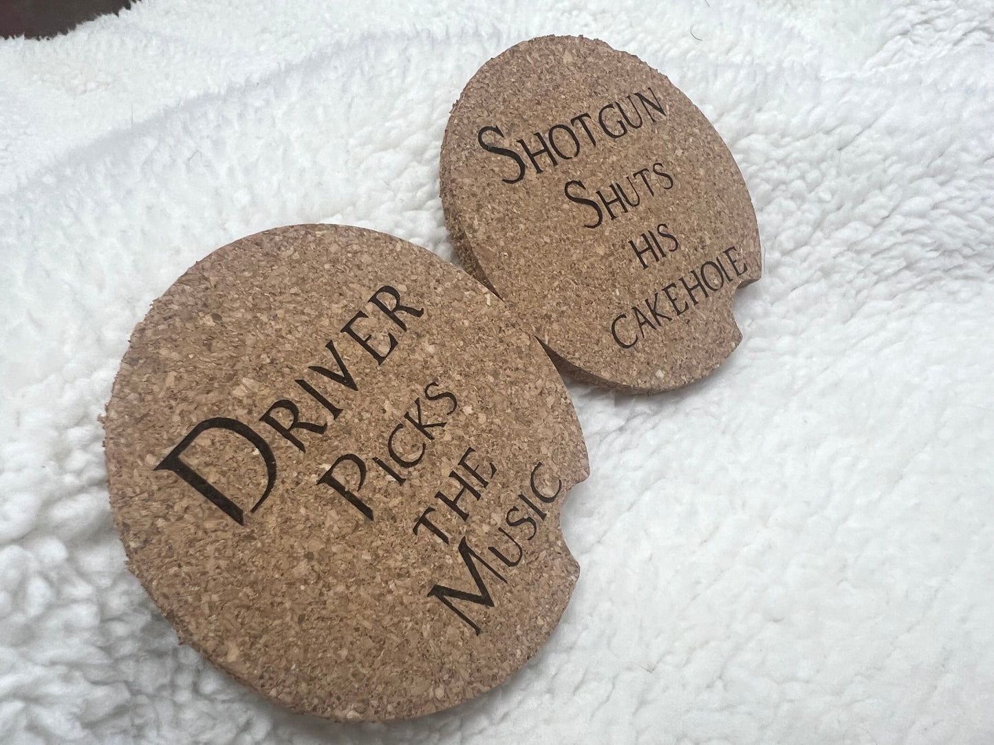 Two MOON AND SKYE CREATIONS LLC cork car coasters rest on a soft white blanket; one says Driver picks the music, while the other adds a personal touch with Shotgun shuts his cakehole.