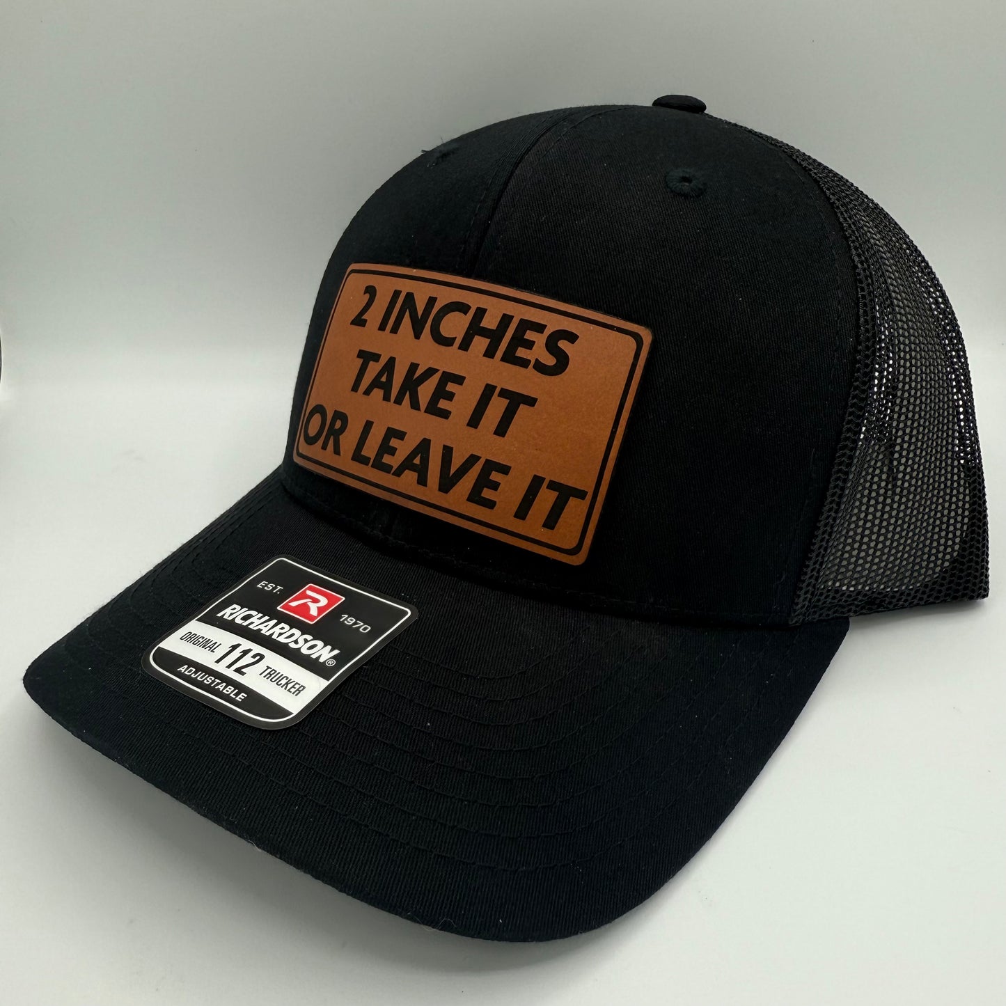 Black Richardson 112 trucker hat with mesh back, featuring a brown leatherette patch engraved with the humorous phrase '2 INCHES TAKE IT OR LEAVE IT' in bold black lettering. A product sticker is visible on the curved brim.