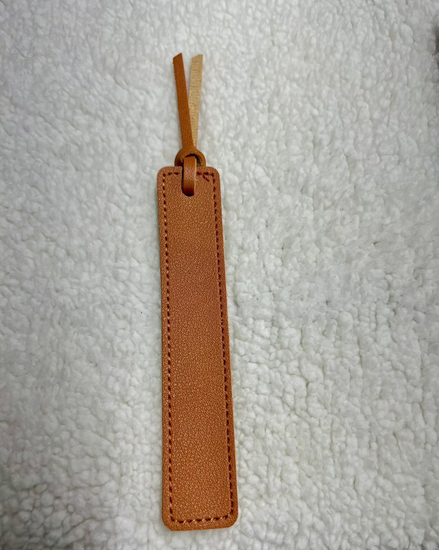 MOON AND SKYE CREATIONS LLCs Leather Bookmarks, designed for book lovers, are brown leather with laser engraving and a looped strap at the top, showcased on a white textured surface.