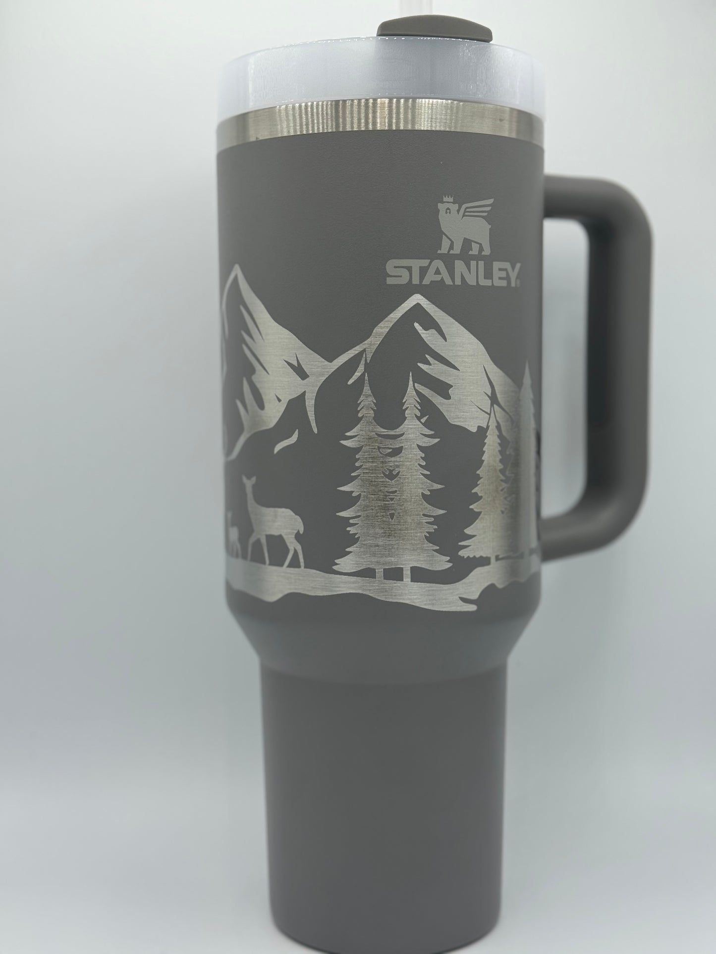 "Custom personalized grey 40oz Stanley tumbler featuring an engraved mountain and forest scene with deer silhouettes. Includes a sturdy handle, a secure lid, and the iconic Stanley logo, perfect for outdoor enthusiasts and adventure lovers."
