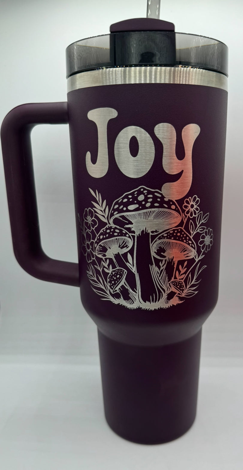 "Custom personalized purple 40oz Stanley tumbler featuring an engraved 'Joy' design with detailed mushrooms and floral elements. Comes with a sturdy handle and a lid with a straw, perfect for keeping beverages hot or cold in style."