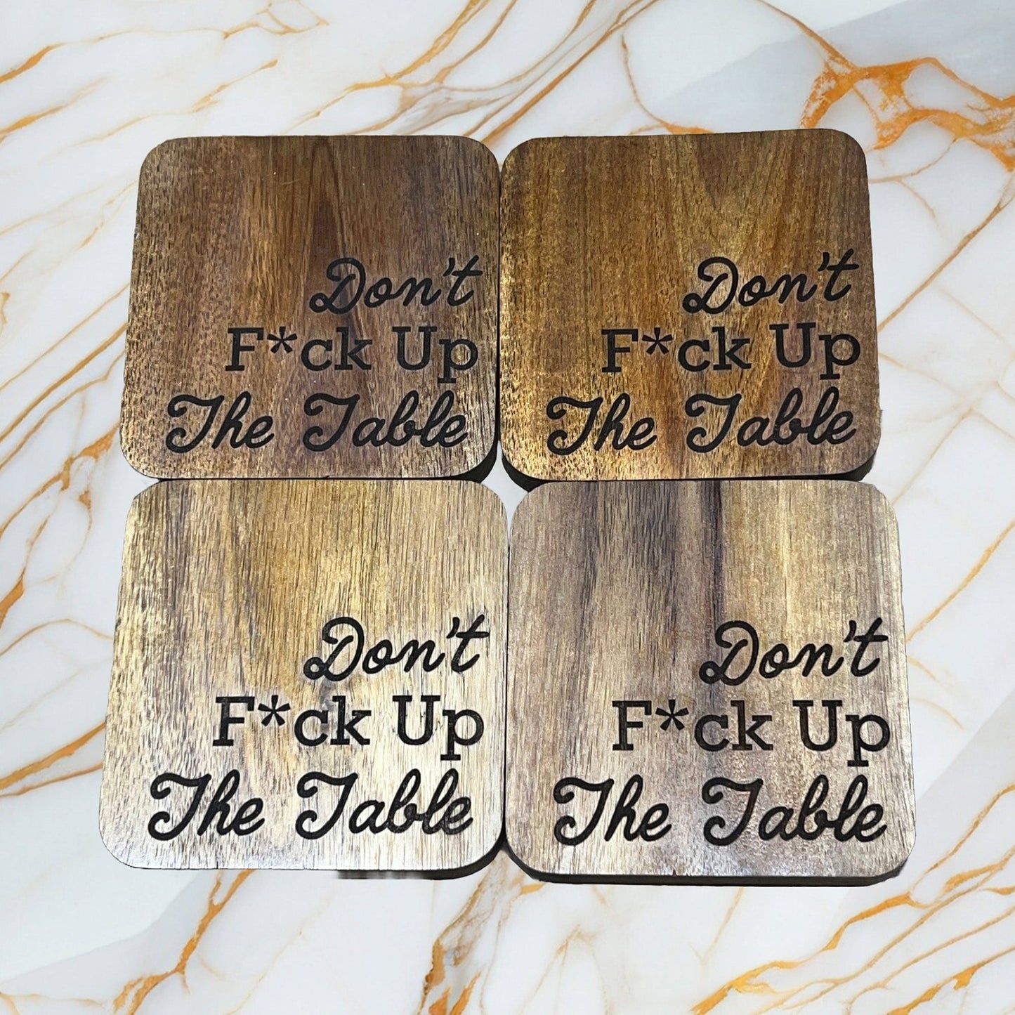 Four wood coasters with the text Dont F*ck Up The Table are arranged in a square on a marble surface. Made by MOON AND SKYE CREATIONS LLC, these coasters feature a distinctive engraved design and polished polyurethane finish, combining style with effective table protection.