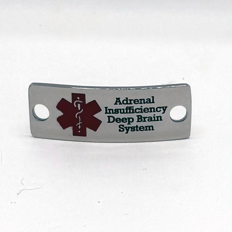 Front view of a polished stainless steel medical alert pendant featuring a red medical symbol on the left side. Engraved text reads 'Adrenal Insufficiency Deep Brain System' in black font. The pendant has a curved shape with two holes for attachment.