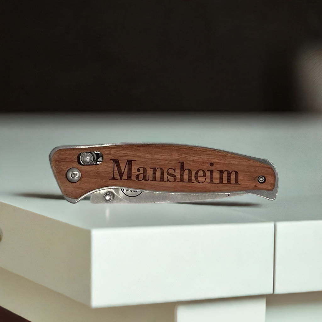 "Personalized engraved Bison River pocket knife featuring a wood handle with the name 'Mansheim' etched in bold serif font. The knife has a sleek stainless steel frame and a compact folding design, resting on a modern white surface."