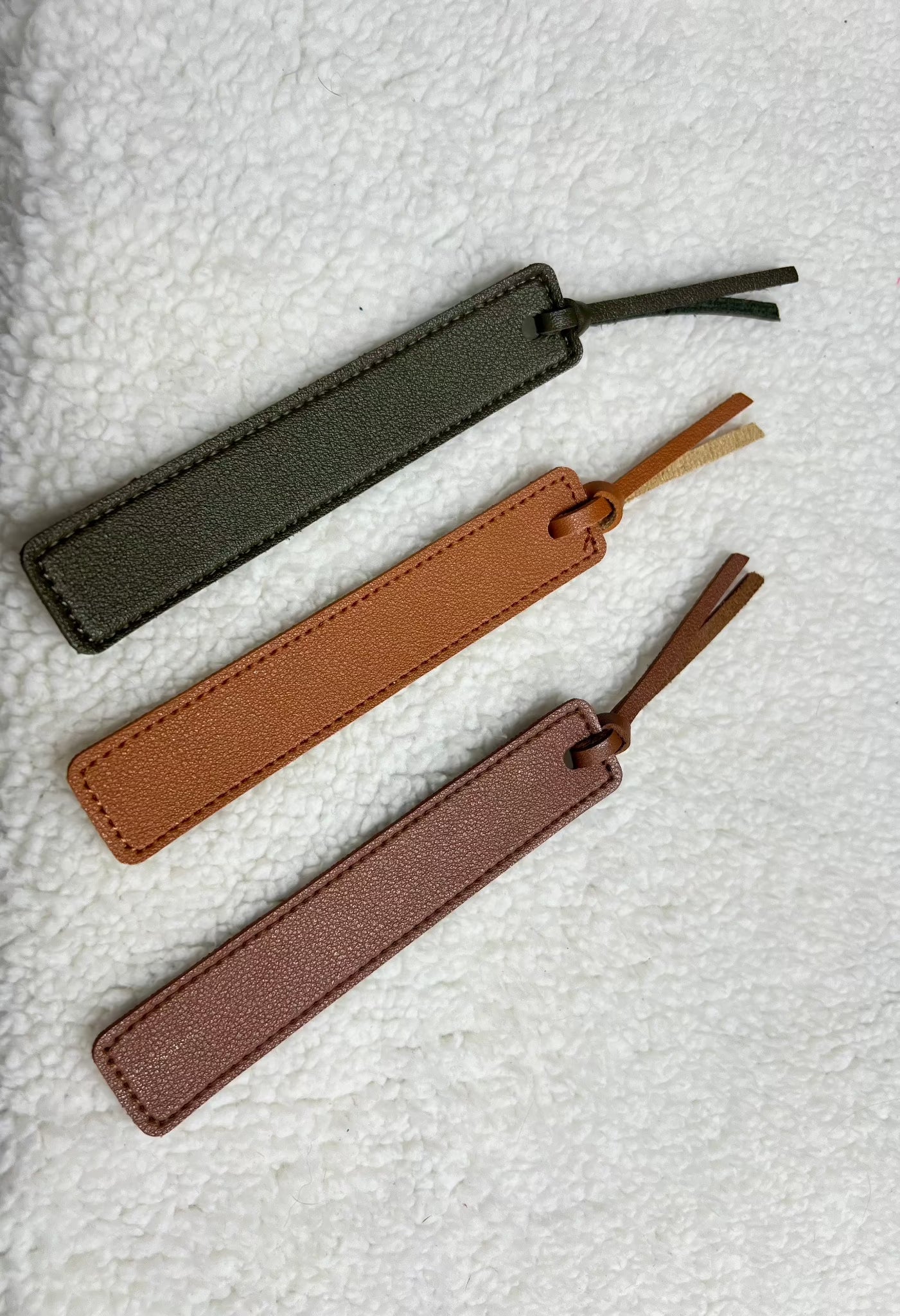 Three MOON AND SKYE CREATIONS LLC rectangular leather bookmarks in olive green, tan, and mauve with matching tassels lie on a textured white surface. Exquisitely laser engraved, these bookmarks are perfect for book lovers seeking an elegant touch.