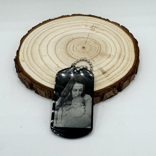 "Custom black stainless steel dog tag keychain or necklace featuring an engraved photo of a mother lovingly embracing her baby, displayed on a rustic wooden background."