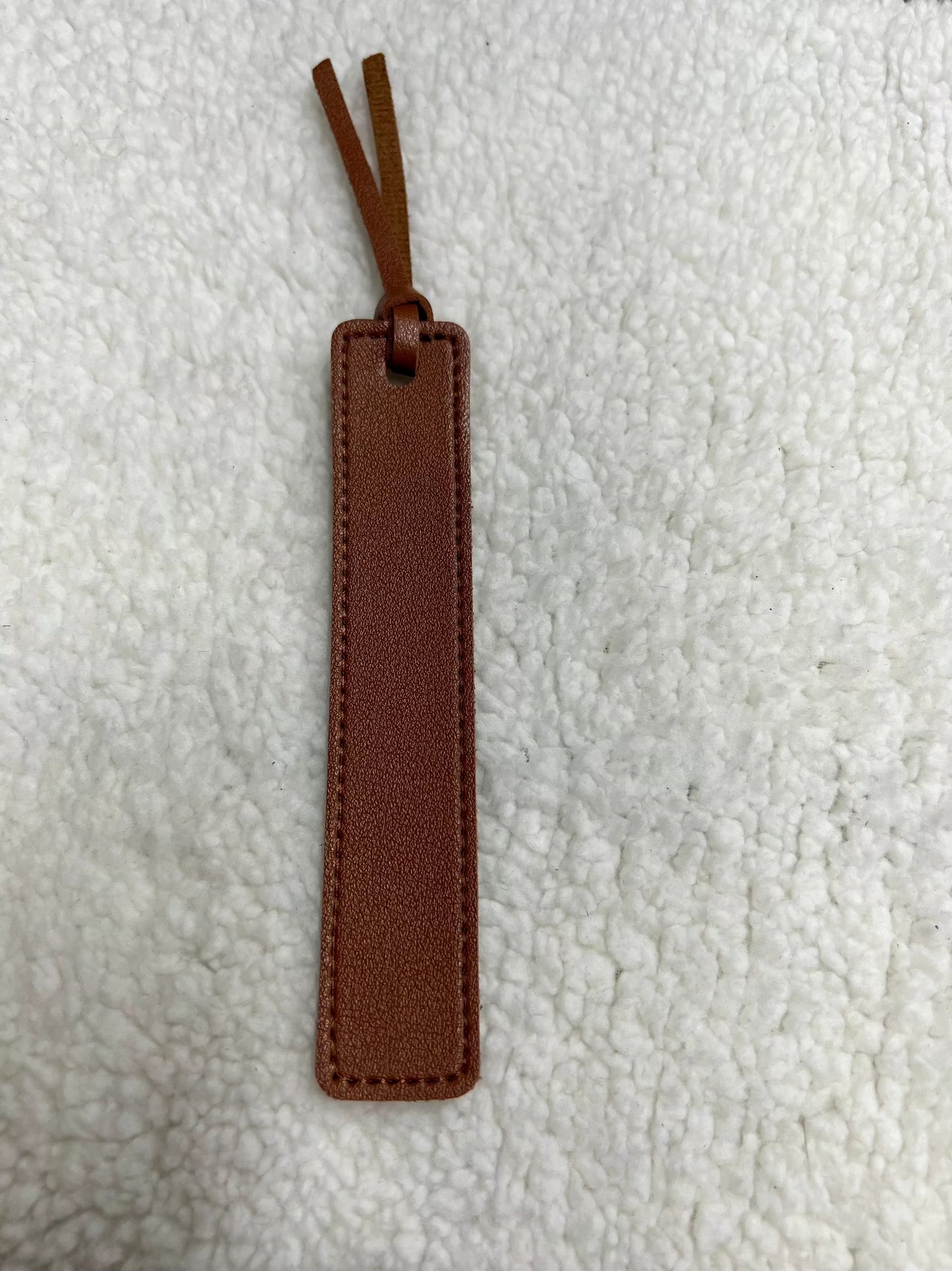 MOON AND SKYE CREATIONS LLCs Leather Bookmark, featuring stitched edges, two leather tassels, and a subtle laser engraved design, is perfect for book lovers. It rests on a textured white blanket.