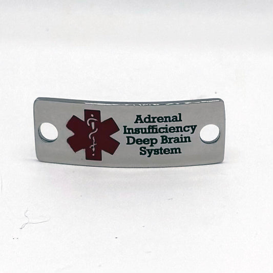 Front view of a polished stainless steel medical alert pendant featuring a red medical symbol on the left side. Engraved text reads 'Adrenal Insufficiency Deep Brain System' in black font. The pendant has a curved shape with two holes for attachment.