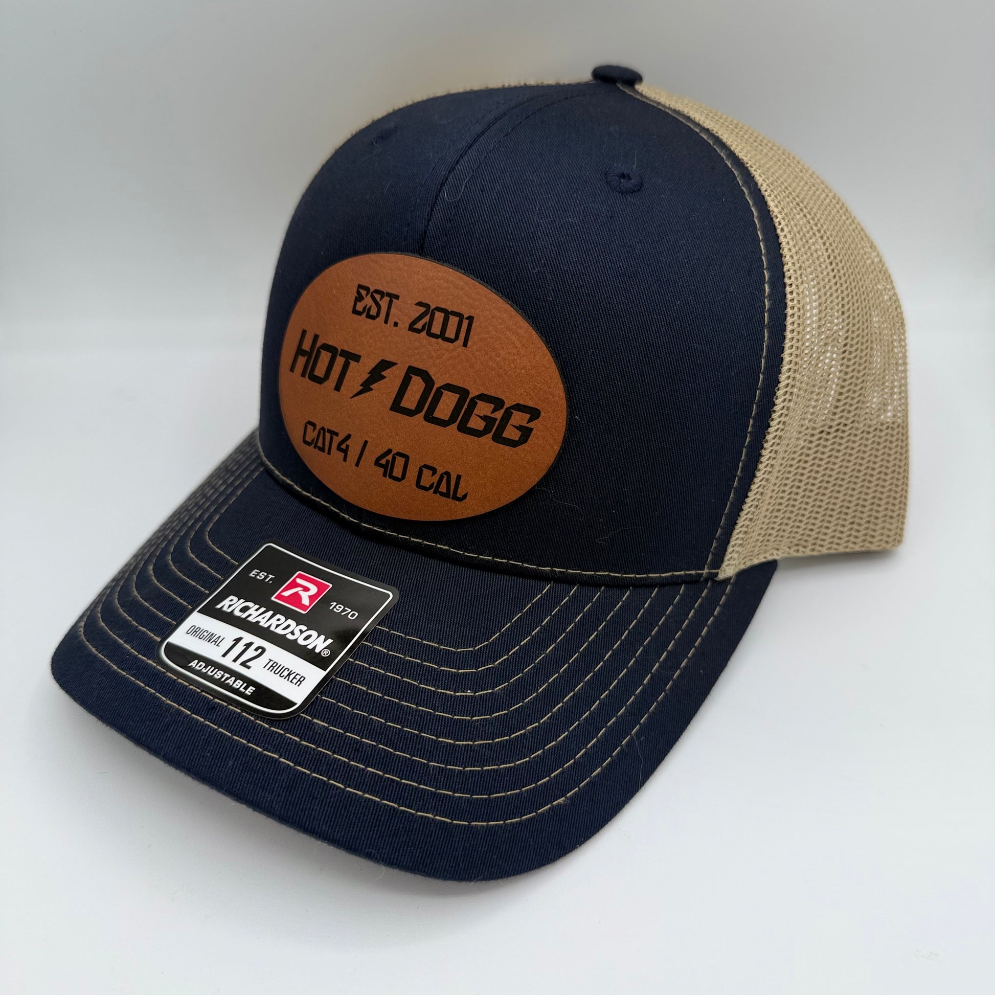 Richardson 112 trucker hat with a navy front, tan mesh back, and curved brim. Features a brown oval leatherette patch engraved with 'EST. 2001 HOT ⚡ DOGG CAT4 / 40 CAL' in bold black text. Manufacturer sticker is visible on the brim.