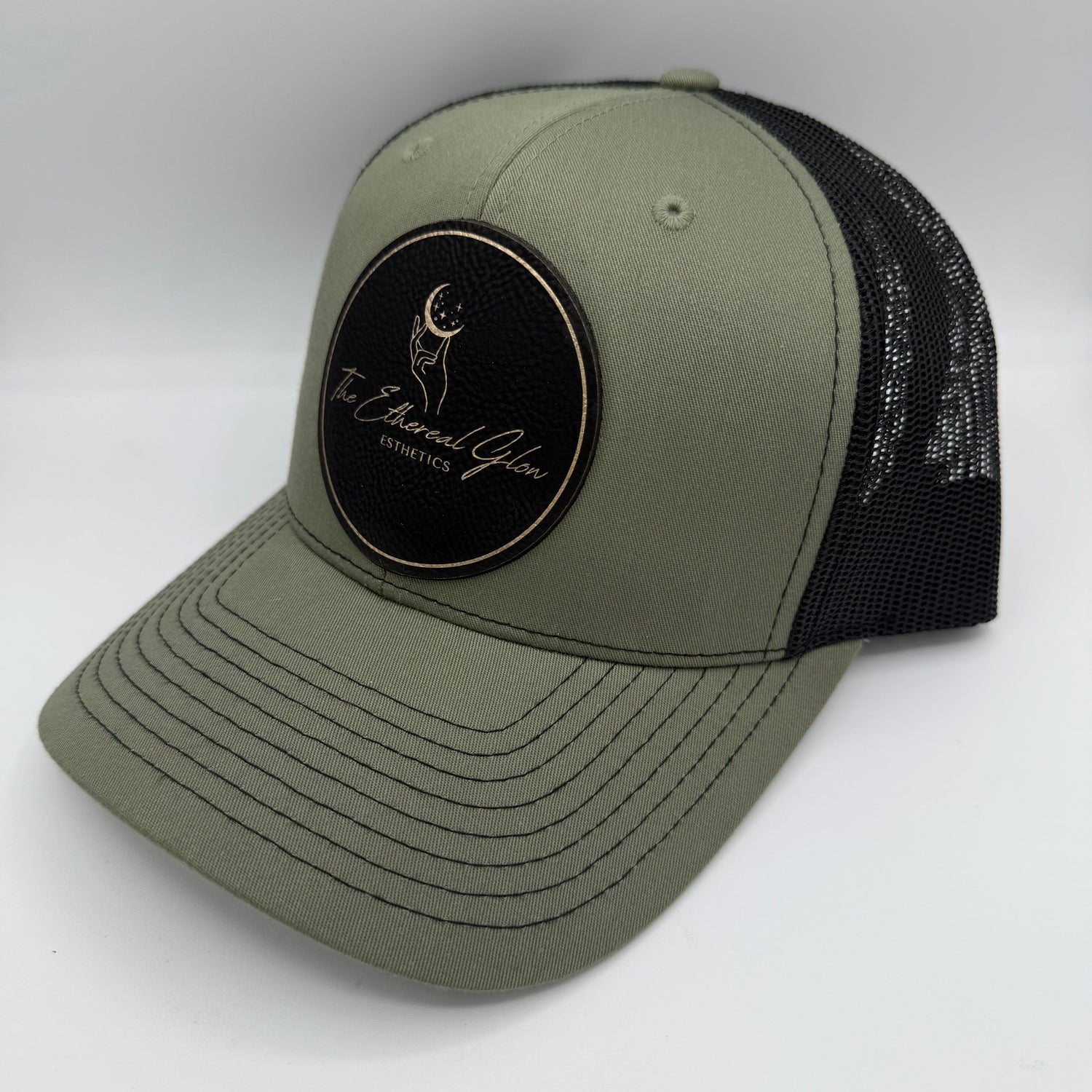 Olive green and black trucker hat featuring a black circular leatherette patch with gold border and gold engraved logo that reads 'The Ethereal Glow Esthetics,' including a hand reaching toward a crescent moon with stars.
