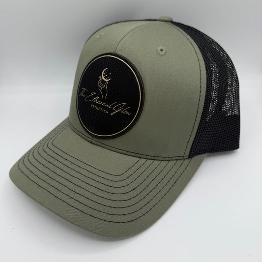 Olive green and black trucker hat featuring a black circular leatherette patch with gold border and gold engraved logo that reads 'The Ethereal Glow Esthetics,' including a hand reaching toward a crescent moon with stars.