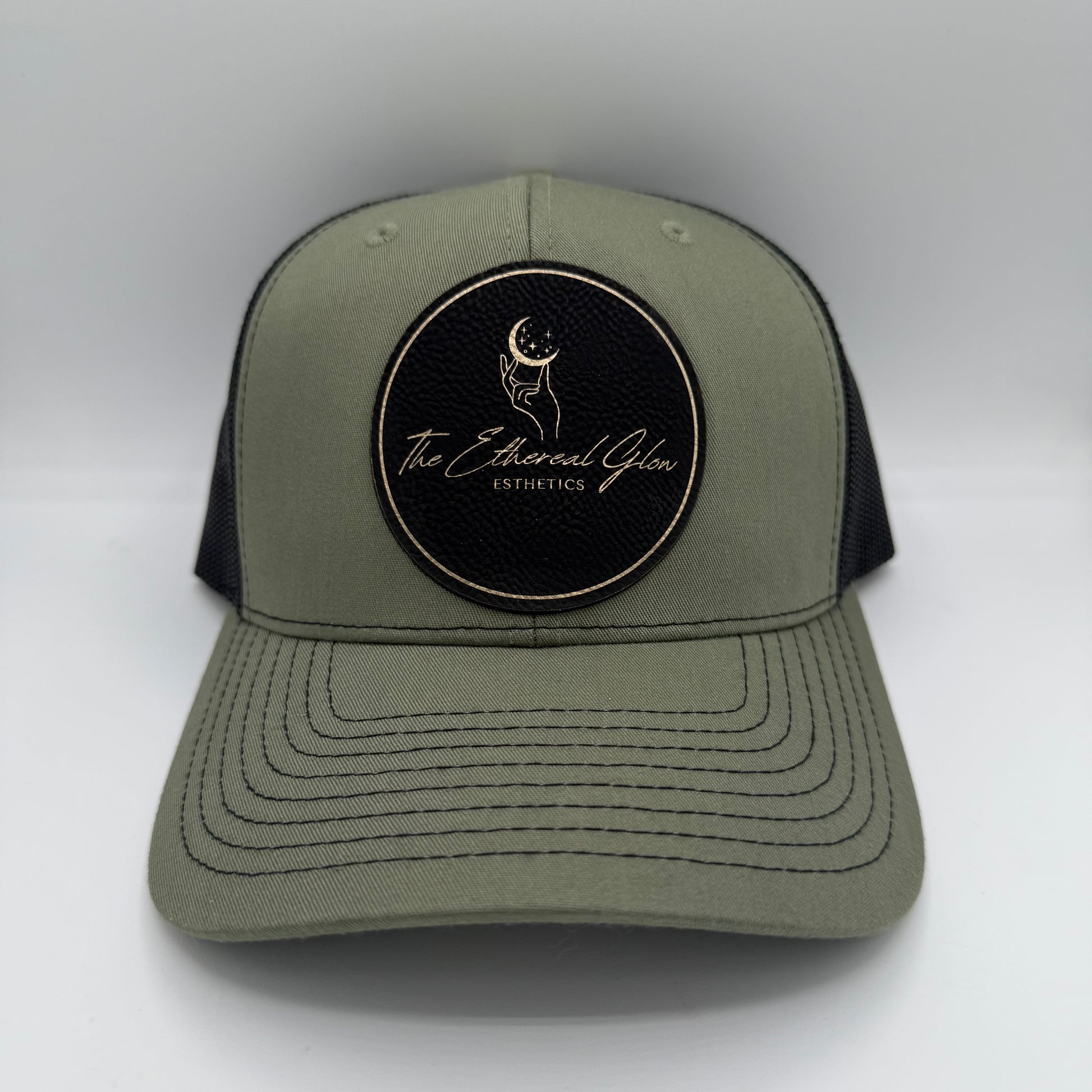 Olive green and black trucker hat featuring a black circular leatherette patch with gold border and gold engraved logo that reads 'The Ethereal Glow Esthetics,' including a hand reaching toward a crescent moon with stars.