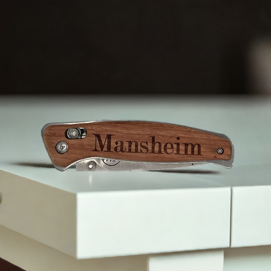 "Personalized engraved Bison River pocket knife featuring a wood handle with the name 'Mansheim' etched in bold serif font. The knife has a sleek stainless steel frame and a compact folding design, resting on a modern white surface."