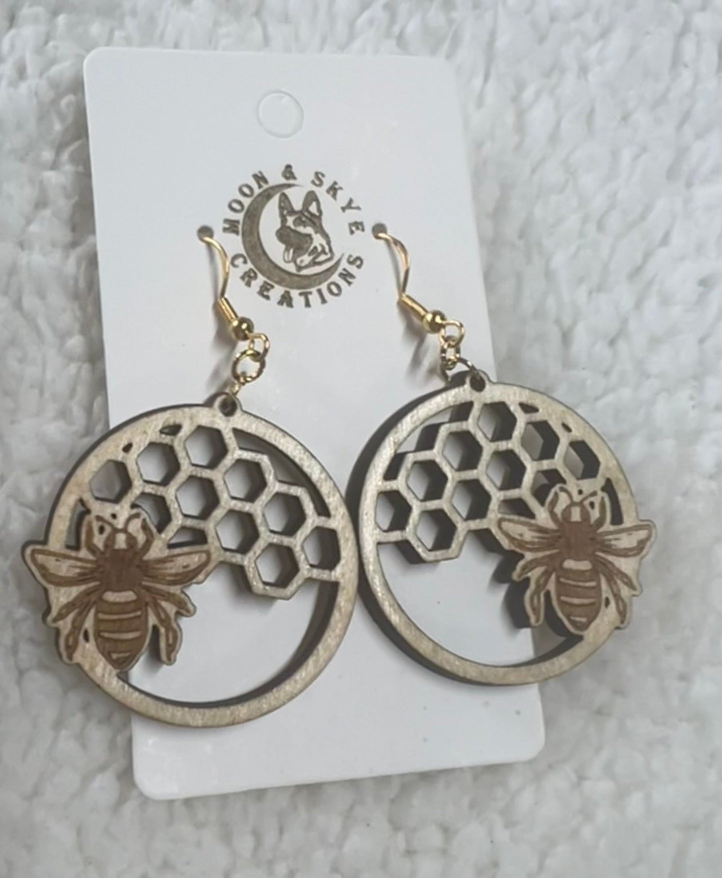 A pair of handcrafted wooden earrings shaped in circular frames featuring a honeycomb cutout pattern with a bee design overlay. The earrings hang from gold hooks and are displayed on a branded card reading "Moon & Skye Creations," set against a soft, white textured background.